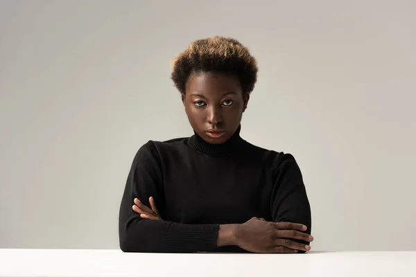 Afro femme au col roulé noir assis à table avec les bras croisés isolés sur gris — Photo de stock