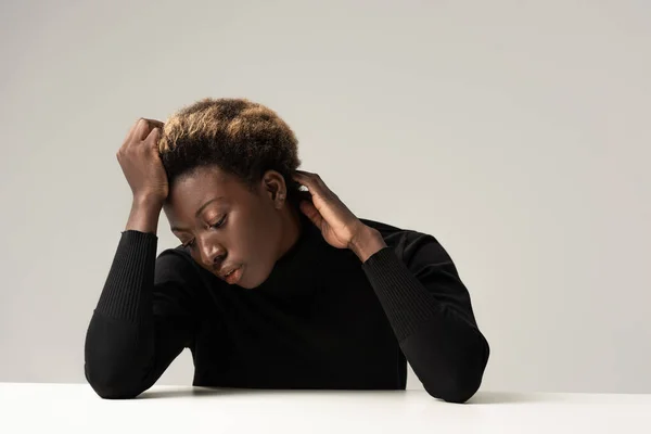 Mujer afroamericana pensativa en cuello alto negro sentada a la mesa aislada en gris - foto de stock