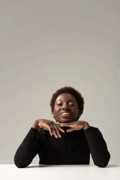 Alegre afroamericana mujer en negro cuello alto sentado a mesa aislado en gris - foto de stock