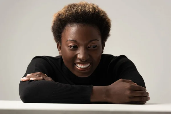 Жизнерадостная африканская американка в черной водолазке, сидящая за столом изолированная на сером — стоковое фото
