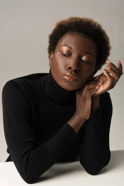 Ніжна афроамериканська жінка в чорному черепасі сидить за столом ізольовано на сірому — стокове фото
