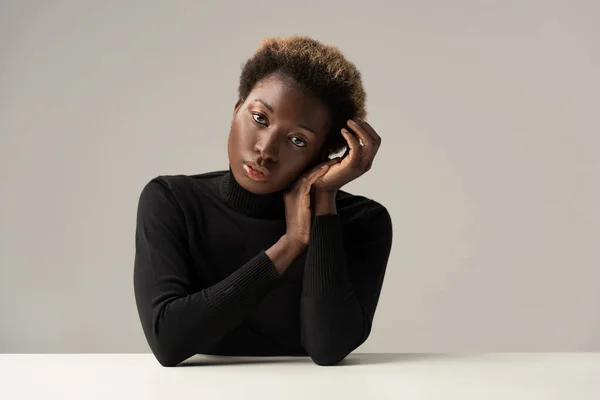 Attractive tender african american girl in black turtleneck sitting at table isolated on grey — Stock Photo