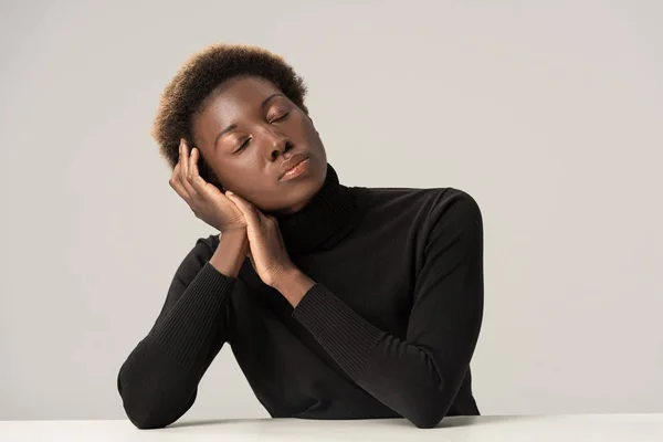 Jolie femme afro-américaine tendre avec les yeux fermés en col roulé noir assis à la table isolé sur gris — Photo de stock