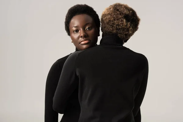 Hermosas mujeres afroamericanas en cuellos altos negros abrazándose aisladas en gris - foto de stock