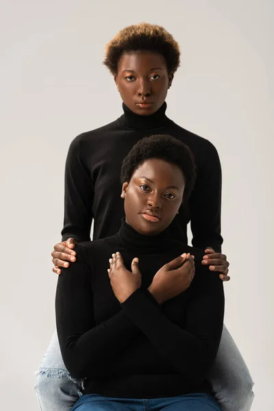 Африканские американские подружки в черных водолазках обнимаются изолированно на серой — стоковое фото