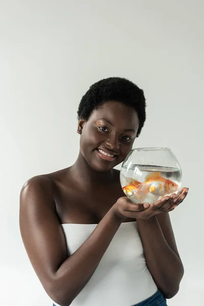 Весела афроамериканська дівчина тримає акваріум з рибою ізольовано на сірому — стокове фото