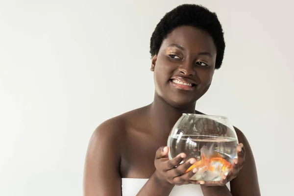 Chica afroamericana sonriente sosteniendo acuario con peces aislados en gris - foto de stock