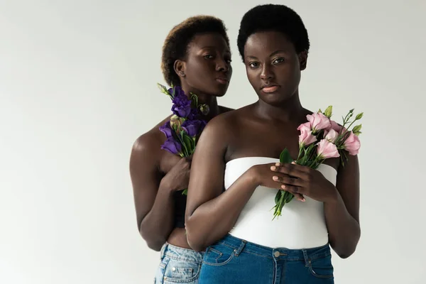 Афроамериканські жінки в джинсах і вершинах тримають квіти ізольовані на сірому — стокове фото