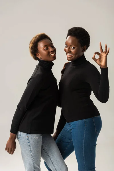 Smiling african american girls in black turtlenecks showing ok sign isolated on grey — Stock Photo