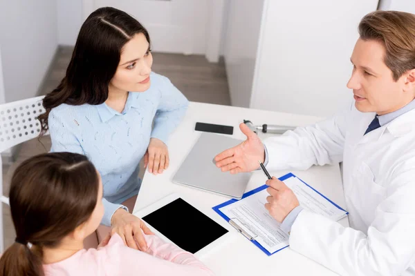 Vista de ángulo alto del otorrinolaringólogo hablando con la madre y la hija cerca de la tableta digital con pantalla en blanco - foto de stock