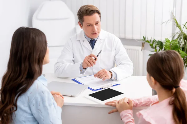 Ernsthafter Arzt im Gespräch mit Mutter und Tochter nahe digitalem Tablet mit leerem Bildschirm auf dem Schreibtisch — Stockfoto