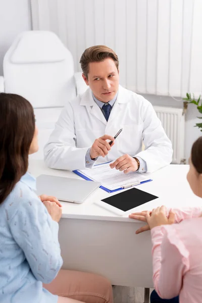 Otorrinolaringólogo serio hablando con la madre y su hija cerca de la tableta digital con pantalla en blanco en el escritorio - foto de stock