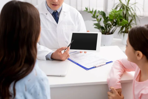 Enfoque selectivo de la mujer con hija cerca de otolaringólogo sonriente mostrando tableta digital con pantalla en blanco - foto de stock