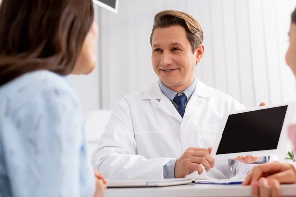 Selective focus of smiling pediatrist showing to mother and daughter digital tablet with blank screen — Stock Photo