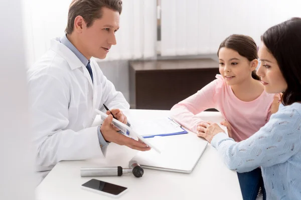 Enfoque selectivo del otorrinolaringólogo mostrando la tabla digital a la madre y a la hija durante la consulta - foto de stock