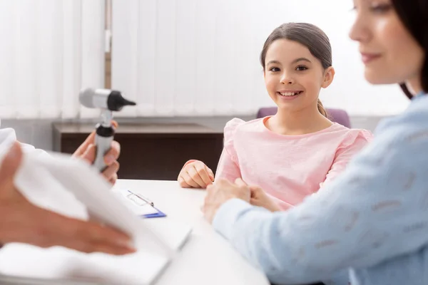 Selective focus of smiling adorable kid sitting at desk near otolaryngologist talking to her mother — Stock Photo