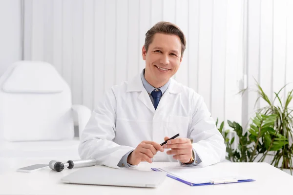 Otorrinolaringólogo sonriente mirando la cámara mientras está sentado en el lugar de trabajo de hormigas cerca de otoscopio, computadora portátil y portapapeles - foto de stock