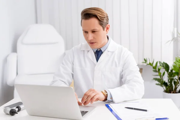 Thoughtful otolaryngologist using laptop at workplace near otoscope and clipboard — Stock Photo