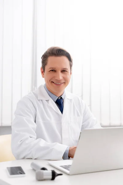 Otorrinolaringólogo sonriente mirando a la cámara mientras usa la computadora portátil en el lugar de trabajo - foto de stock