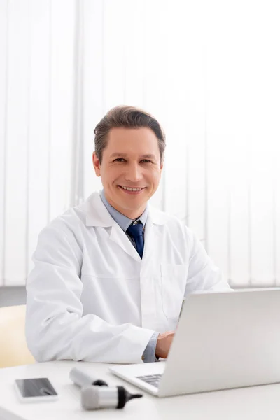 Otorrinolaringologista sorrindo olhando para a câmera enquanto usa laptop no local de trabalho — Fotografia de Stock