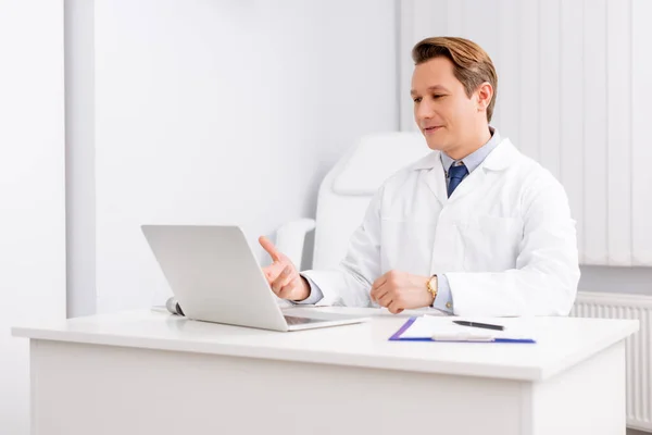 Handsome otolaryngologist having video chat on laptop while sitting at workplace — Stock Photo