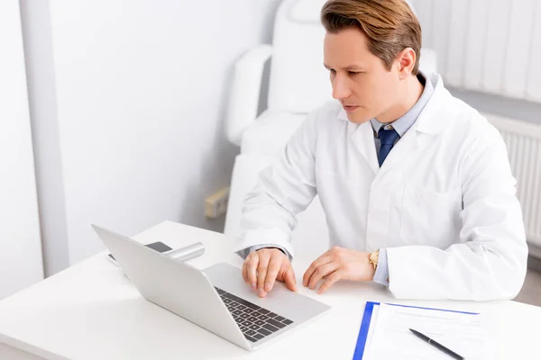 Médico atento usando el ordenador portátil mientras está sentado en el lugar de trabajo — Stock Photo