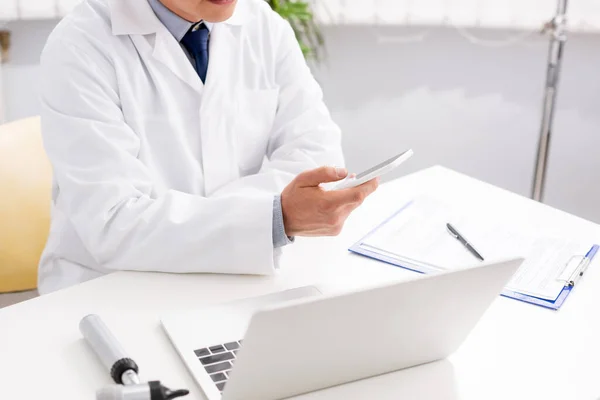 Cropped view of otolaryngologist using smartphone near clipboard, laptop and otoscope — Stock Photo