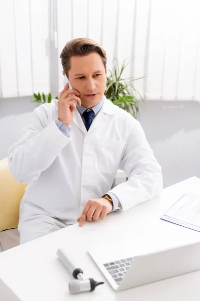 Otorrinolaringólogo guapo hablando en el teléfono inteligente mientras está sentado en el lugar de trabajo cerca de portátil y otoscopio — Stock Photo