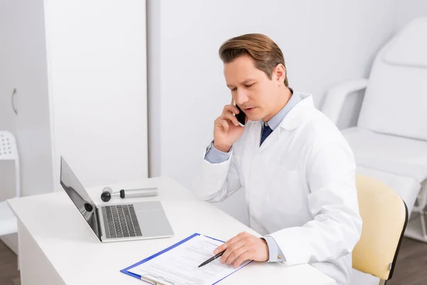 Médico serio hablando en el teléfono inteligente mientras está sentado en el lugar de trabajo cerca de portapapeles, portátil y otoscopio - foto de stock