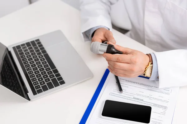 Vista recortada del otorrinolaringólogo que sostiene el otoscopio cerca del portapapeles, computadora portátil y teléfono inteligente con pantalla en blanco - foto de stock