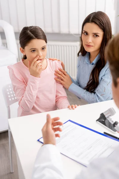 Vista recortada de ent médico, y mujer calmando hija tocándose la nariz - foto de stock