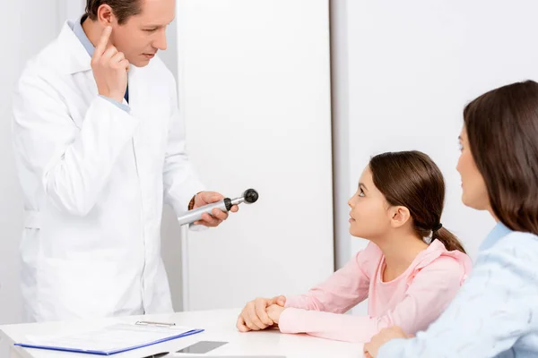 Madre e hija mirando al otorrinolaringólogo sosteniendo otoscopio y señalando con el dedo su oreja - foto de stock