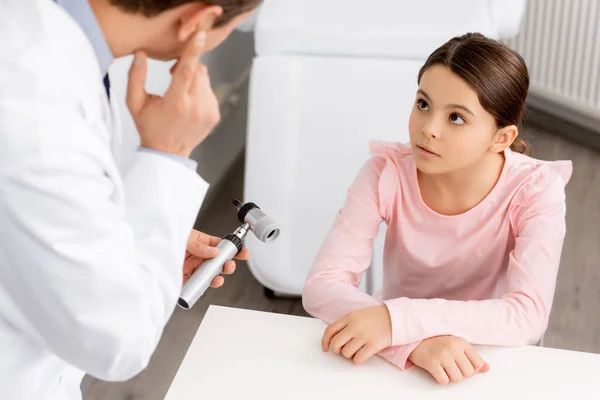 Cropped view of otolaryngologist pointing at his ear while showing otoscope to attentive adorable child — Stock Photo