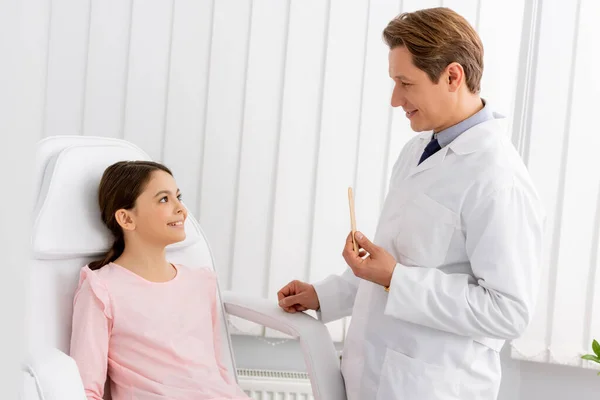 Otorinolaringoiatra sorridente che tiene la lingua depressore mentre in piedi vicino a bambino allegro seduto sulla sedia medica — Foto stock