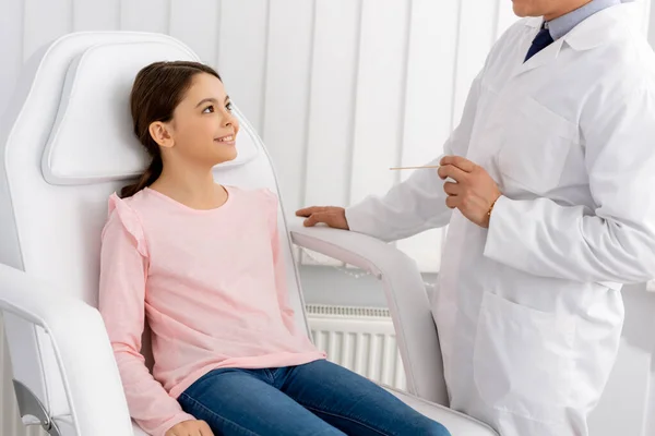 Cropped view of otolaryngologist holding throat spatula while standing near smiling kid sitting in medical chair — Stock Photo