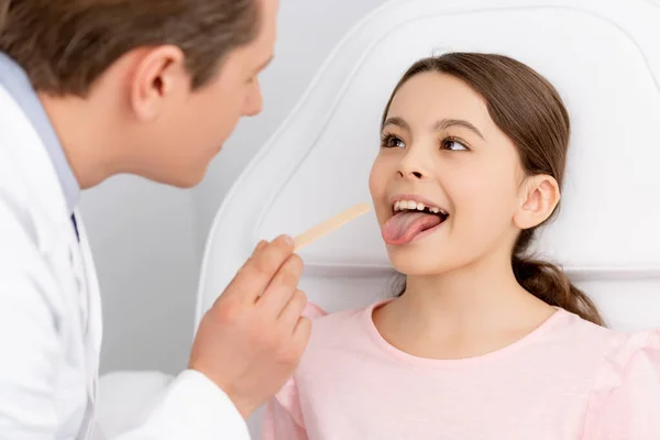 Enfoque selectivo de ent médico sosteniendo depresor lengua cerca lindo niño sobresaliendo lengua - foto de stock