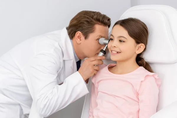 Attentive ent physician examining ear of smiling kid with otoscope — Stock Photo