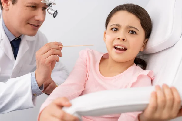 Sonriente ent médico celebración lengua depresor cerca asustado niño - foto de stock