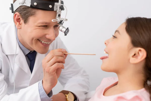 Cheerful ent physician examining throat of cute child with tongue depressor — Stock Photo