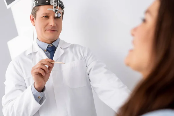 Selective focus of handsome otolaryngologist in ent headlight holding throat spatula near patient — Stock Photo