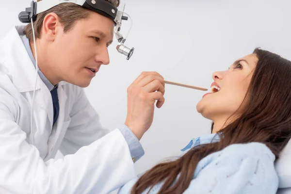 Attentive otolaryngologist in ent headlight examining throat ao attractive patient with throat spatula — Stock Photo