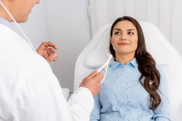 Visão parcial do otorrinolaringologista segurando espéculo nasal perto do atraente paciente sorridente sentado em cadeira médica — Fotografia de Stock