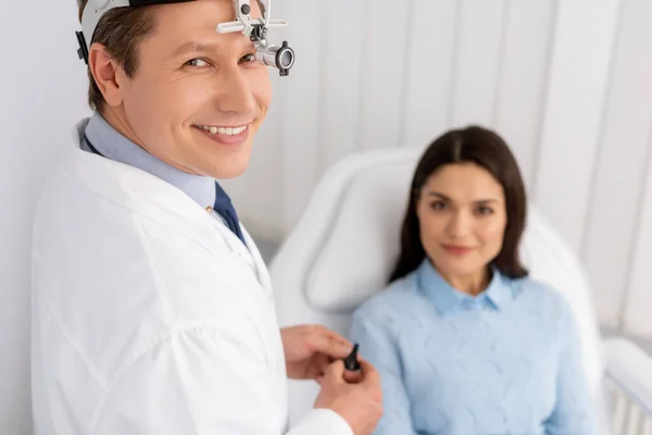 Enfoque selectivo del otorrinolaringólogo sonriente en el faro de ent que sostiene el espéculo del oído cerca de la mujer sonriente sentada en la silla médica - foto de stock