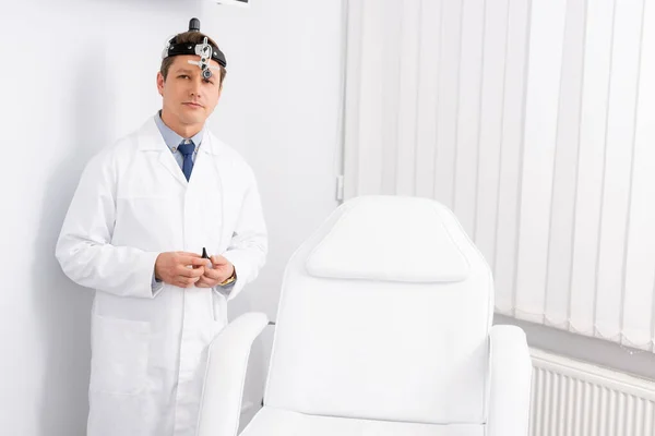 Schöner, selbstbewusster HNO-Arzt mit Stirnlampe, der in der Nähe des Medizinstuhls steht und in die Kamera blickt — Stockfoto
