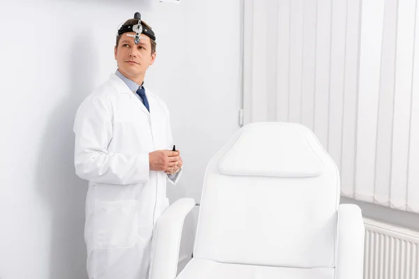 Bonito, otorrinolaringologista confiante com farol ent em pé perto da cadeira médica — Fotografia de Stock