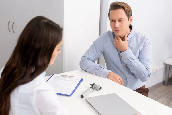 Handsome man touching throat while sitting near otolaryngologist in clinic — Stock Photo