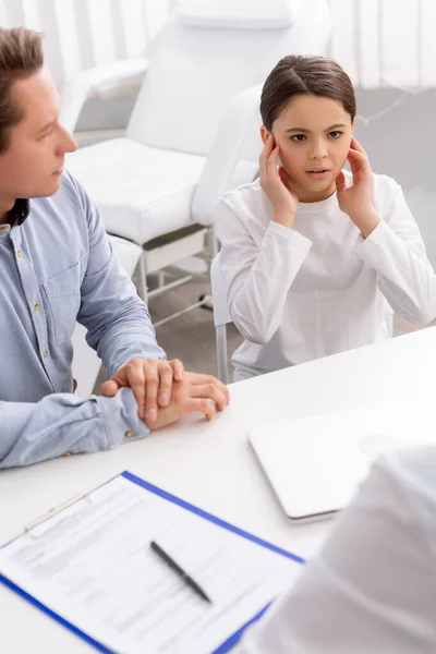 Worried kid touching ears while sitting with father on consultation with ent physician — Stock Photo