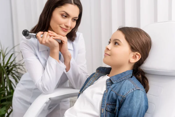 Un medico sorridente che tiene l'otoscopio mentre osserva il bambino adorabile seduto sulla sedia medica — Foto stock