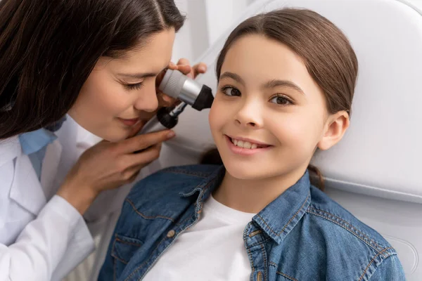 Внимательный отоларинголог осматривает ухо улыбающегося ребенка с помощью отоскопа — стоковое фото