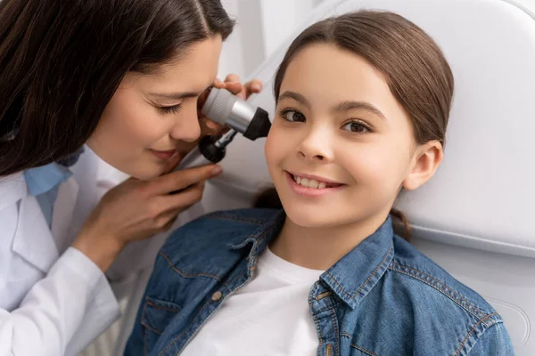 Attentive otolaryngologist examining ear of smiling child with otoscope — Stock Photo
